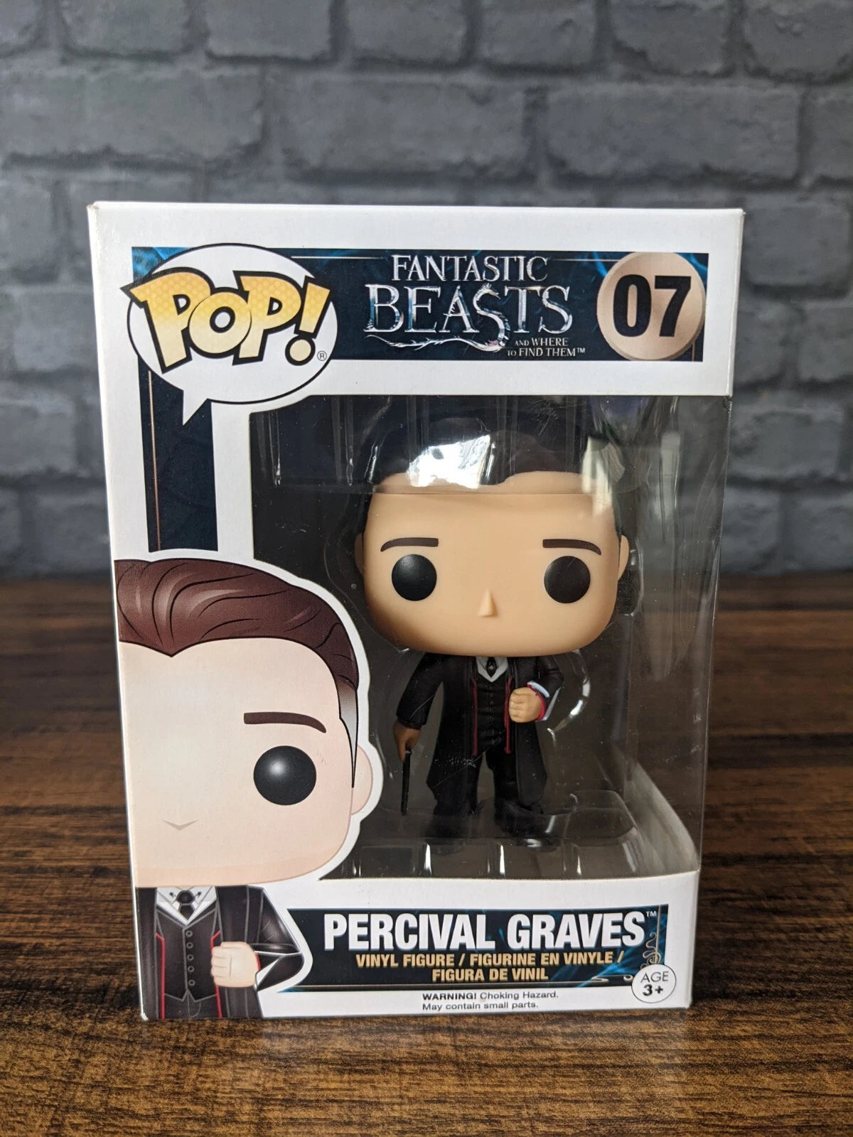 Funko Pop! vinyl figure of Percival Graves from Fantastic Beasts, number 07, in its original window box packaging, displayed on a wooden surface against a gray brick wall.