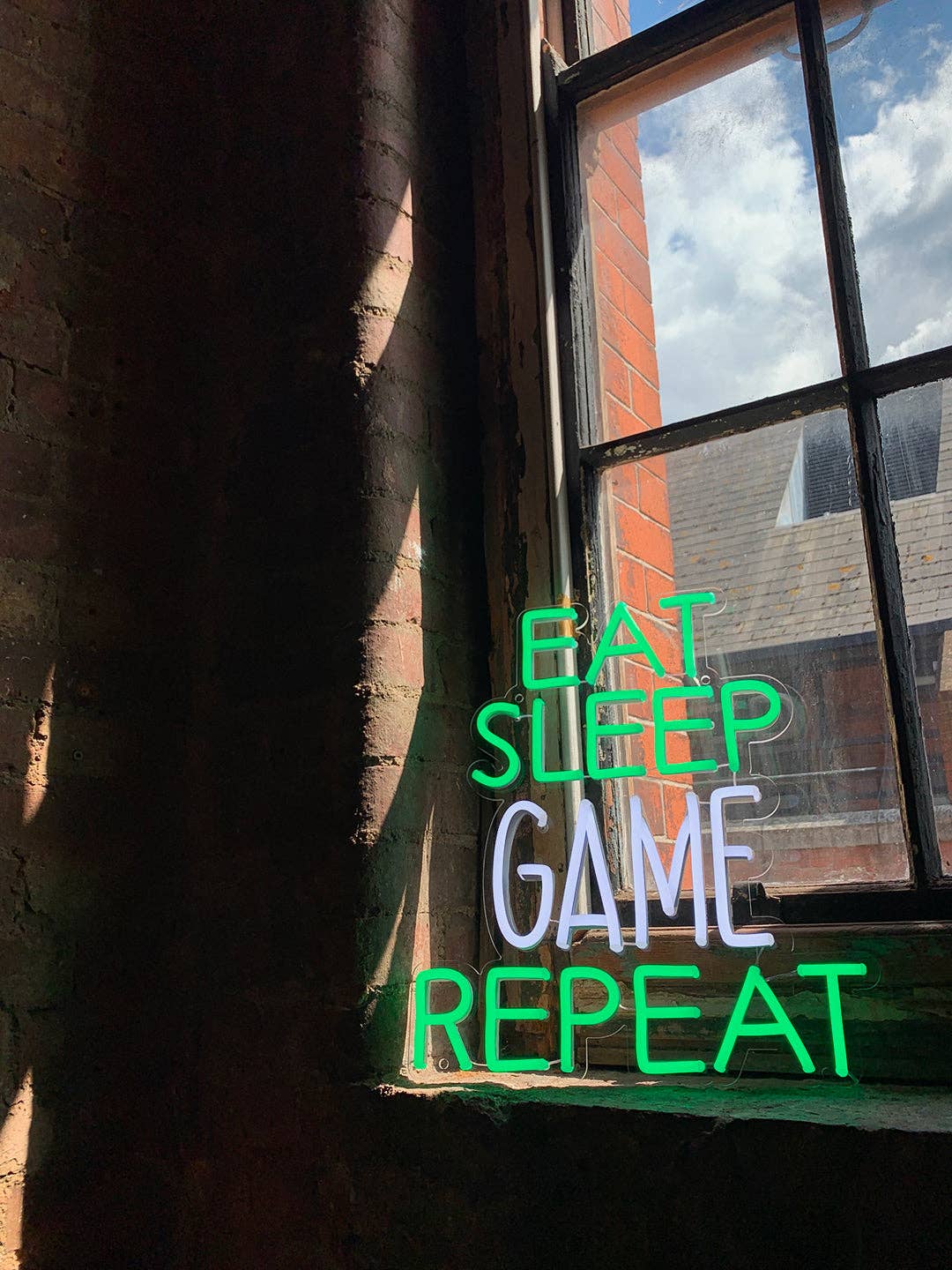 Neon sign with 'Eat Sleep Game Repeat' text in front of a window with a view of a building.