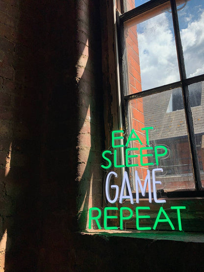 Neon sign with 'Eat Sleep Game Repeat' text in front of a window with a view of a building.