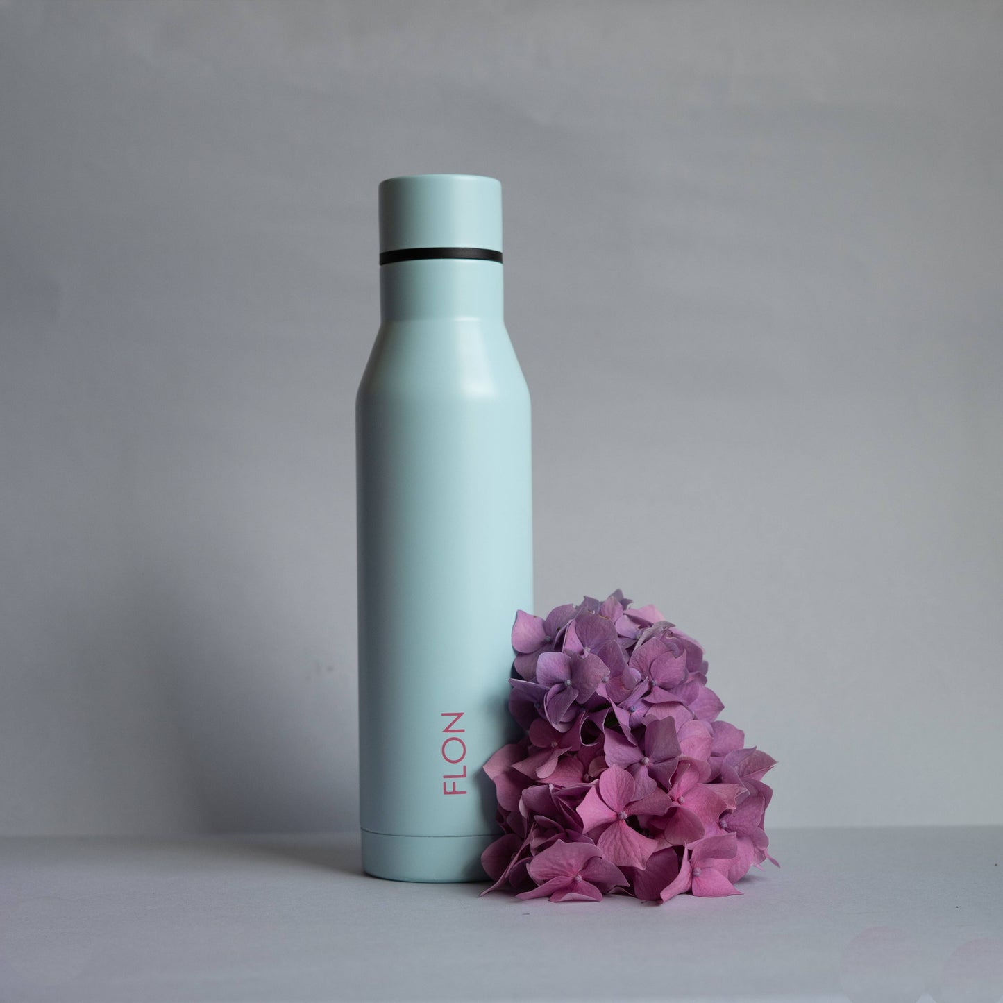 Light blue water bottle with 'FLO' branding next to pink flowers on a gray background