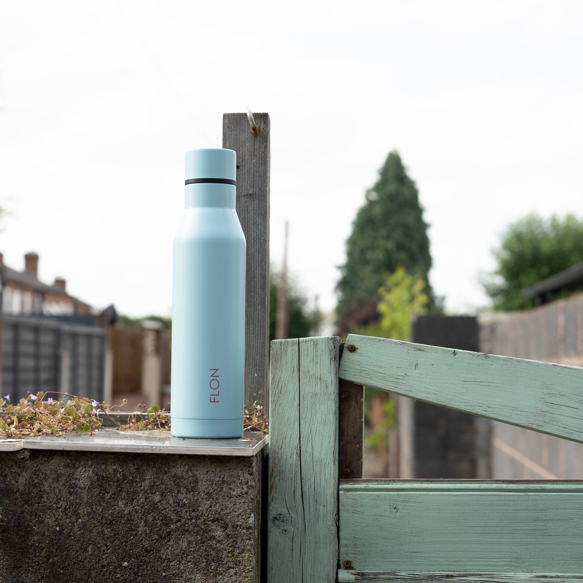 Light blue water bottle with 'FLON' branding on a wooden fence outdoors.